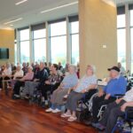 Stranski pogled na starejše občinstvo, ki sedi v dvorani z velikimi okni, nekateri na invalidskih vozičkih, z nasmejanimi izrazi med spremljanjem dogajanja.