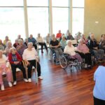 Skupina starejših ljudi, ki sedijo v dvorani z lesenimi tlemi in velikimi okni, obrnjeni proti nastopajočim. Nekateri izmed njih so v invalidskih vozičkih, medtem ko spremljajo dogajanje.