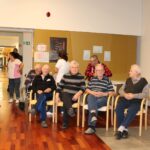 Skupina starejših moških sedi v vrsti na stolih v skupnem prostoru doma za starejše. V ozadju so vidni zaposleni in obiskovalci, ki se zbirajo okoli. Nekateri starejši moški so v invalidskih vozičkih, drugi sedijo na stolih in sproščeno čakajo na dogodek.
