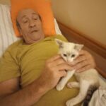 Starejši moški leži v postelji, naslonjen na oranžno blazino. V rokah drži majhnega mucka, ki mu sedi na prsih. Moški ga nežno boža, ob tem pa je videti umirjen in zadovoljen. Prostor je osvetljen z mehko svetlobo, vzdušje pa deluje domače.