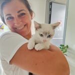 Nasmejana ženska v beli uniformi drži v rokah majhnega mucka. Muc se zdi sproščen, z zanimanjem zre naravnost v kamero. Ženska ga tesno stiska k sebi, oba pa izžarevata toplino in navezanost. Prostor v ozadju je svetel, z vrati in nekaj lončnicami.