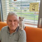 Starejši moški sedi na oranžnem kavču z majhnim mucom, ki mu sedi na rami. Oba gledata naravnost v kamero. V ozadju so okna z žaluzijami, skozi katera se vidi zelenje zunaj. Na oknu je tudi znak z napisom "Ne odpiraj okna na kip!!" in risba črnega mačka.