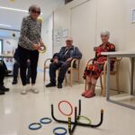 Starejša ženska, oblečena v črno-belo majico z zebrastega vzorca, sodeluje v igri metanja obročev. Sklonjena je naprej, pripravljena, da vrže obroč proti tarči na tleh. Ob njej sedita dva starejša udeleženca, eden v temni jopici in s kapo, drugi pa v pisani obleki. Očitno uživajo v dejavnosti v svetlem prostoru, kjer so obešene dekorativne sončnice.