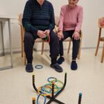 Starejši moški in ženska sedita na stolih in gledata igro metanja obročev. Moški drži nekaj obročev v rokah in se pripravlja, da jih vrže, medtem ko na tleh pred njim leži več obročev, ki so pristali okoli tarče. Videti je, da se oba osredotočata na igro, v prostoru pa vlada sproščeno vzdušje.
