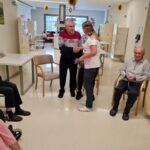 Starejši moški v rdeče-črtasti majici stoji in se rokuje z negovalko, ki je oblečena v belo uniformo. Oba sta nasmejana, medtem ko drugi udeleženci sedijo in opazujejo dogajanje. Prostor je svetel in urejen, z nekaj mizami in stoli, kar ustvarja prijetno in domačno vzdušje.