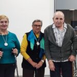 Skupina petih starejših oseb, trije moški in dve ženski, stoji pred svetlim ozadjem in se nasmiha. Vsak ima okoli vratu medaljo. Eden od moških drži priznanje za osvojeno drugo mesto na tekmovanju. Vzdušje je veselo, vsi so urejeni in ponosni na svoje dosežke.