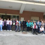 Velika skupina ljudi, sestavljena iz starejših udeležencev in mlajših spremljevalcev, stoji pred vhodom v stavbo. Nekateri udeleženci so na invalidskih vozičkih, drugi stojijo s pripomočki za hojo. Vzdušje je sproščeno in prijateljsko, skupina je pripravljena na skupinsko fotografijo.