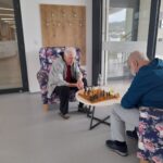 Dva starejša moška sedita na udobnih naslanjačih z rožastim vzorcem in igrata partijo šaha. Okolje je svetlo, moderna zasnova prostora pa vključuje veliko okno, ki omogoča pogled ven. Moška sta globoko osredotočena na igro.