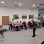 Skupina starejših ljudi se udeležuje športnega dogodka v zaprtem prostoru. Moški na sprednjem delu slike meče žogo v koš, medtem ko ga opazujejo trije sodniki za mizo in občinstvo v ozadju. Vzdušje je sproščeno in zabavno, vsi udeleženci se zabavajo.