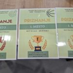 Na leseni mizi so prikazana tri priznanja, vsako z ikono pokala. Priznanja so v slovenščini z naslovom "PRIZNANJE" in prikazujejo naslednje nagrade: tretje mesto za skupinsko metanje na tarčo, prvo mesto za skupinsko metanje na koš ter prvo mesto za individualno metanje na koš.