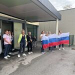 Skupina žensk stoji pred stavbo in drži veliko zastavo v barvah slovenske zastave. Ženske so videti kot osebje ali udeleženke dogodka. Nekatere stojijo pri vhodu v stavbo, druge pozirajo z zastavo. Vzdušje je živahno, saj se zdi, da skupina praznuje ali izkazuje narodni ponos.