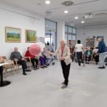 Skupina starejših ljudi sedi ob steni v svetlem prostoru, z žensko v bež jakni, ki stoji na sredini in drži velik roza balon. V ozadju se zdi, da je druga skupina ljudi vključena v dejavnosti; ena oseba stoji, drugi sedijo. Prostor ima velika okna, slike na stenah in sodobno, minimalistično zasnovo.