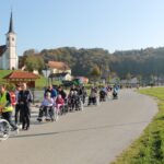 Procesija ljudi v invalidskih vozičkih, ki jih spremljevalci potiskajo po cesti, vodi mimo cerkve na hribu. V ozadju je gozdnato pobočje.