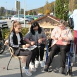 Skupina ljudi sedi na prostem, dve osebi v invalidskih vozičkih, dve pa na stolih. Ukvarjajo se z luščenjem kostanja, v ozadju so parkirani avtomobili in manjša ograja.
