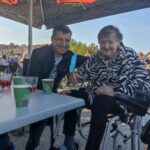 Moški in starejša ženska, oblečena v črno-beli jopič z živalskim vzorcem, sedita za mizo na prostem. Nasmehnjena ženska drži majhno papirnato rožico, na mizi so kozarci z rdečo pijačo in skodelice.