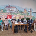 Skupina starejših občanov sedi za mizo pred stensko poslikavo, ki prikazuje mesto z značilnimi stavbami in hribi v ozadju. Na mizi so dokumenti in dekorativni aranžma.