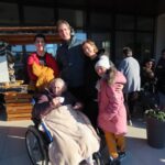 Na sliki je družina, ki pozira ob starejši gospe, ki sedi na invalidskem vozičku z odejo in skodelico v roki. V ozadju so drugi stanovalci doma za starejše, ki uživajo na sončnem dnevu. Ob strani je lesena, praznično okrašena stojnica.