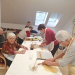 Starejši moški in ženske sodelujejo pri pripravi božičnih dobrot. Nekateri sedijo in uživajo v druženju, medtem ko drugi na mizi valjajo testo. Prostorna soba ima veliko naravne svetlobe, ki vstopa skozi strešna okna.