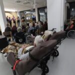 Pogled iz občinstva. V ospredju so starejši obiskovalci, nekateri na invalidskih vozičkih ali v ležalnikih, pokriti z odejami. V ozadju na odru nastopa pevska skupina. Prostor je svetel, okrašen z božičnimi okraski in snežinkami.