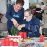 Gospa v modrem puloverju pomaga gospodu, ki v rokah drži zelenje in pripravo za adventni venček. Na mizi so rdeče in bele sveče, ki čakajo na postavitev.