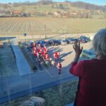 Fotografija prikazuje pogled skozi okno doma za starejše občane. Na dvorišču je skupina ljudi, oblečenih v kostume Božičkov, zbranih ob motorjih in kolesih. V ospredju, v notranjosti, starejša oseba maha proti skupini zunaj. V ozadju je podeželska pokrajina z njivami in griči.