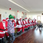 Fotografija prikazuje večjo skupino ljudi, oblečenih v kostume Božičkov, ki stoji v vrsti v domu za starejše. V ospredju je oseba z zeleno božično kapo, ki nekaj pripoveduje, medtem ko stanovalci sedijo in spremljajo dogajanje. Prostor je praznično okrašen.