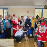 Druga fotografija prikazuje skupinsko sliko. Skupina ljudi različnih starosti, med njimi tudi Božiček in pomočnica v rdeči obleki s kapo, stoji in sedi skupaj v sobi. Večina udeležencev ima v rokah darilne vrečke, nekateri se smehljajo in mahajo v kamero. Ob njih je tudi manjši pes.