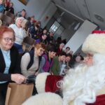 Starejša gospa, ki sedi v prvi vrsti, prejema darilo od Božička, oblečenega v rdečo obleko z belo brado. V ozadju ostali stanovalci z zanimanjem opazujejo dogajanje, nekateri fotografirajo trenutek.
