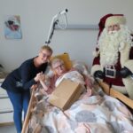 Božiček stoji ob postelji, kjer leži starejša gospa, ki drži rjavo darilno vrečko. Poleg nje stoji ženska, ki se z nasmeškom drži njene roke. Prostor je domače urejen z zelenimi lončnicami in dekoracijami.