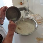 Detajl priprave testa. Ženske roke mešajo sestavine v veliki kovinski posodi. Druga roka drži lonec, iz katerega vliva tekočino v posodo. V ozadju je kuhinjski pult s kuhinjsko krpo in umivalnikom.
