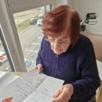 Starejša gospa v modrem puloverju sedi za mizo ob oknu v domu za starejše občane. V rokah drži ročno napisano pismo, ki ga prebere. V ozadju je viden pogled na parkirišče in okoliško podeželje.