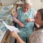 Na fotografiji je starejša gospa v cvetličnem predpasniku, ki sedi na invalidskem vozičku ob svoji postelji. Ob njej je zaposlena, ki ji pomaga brati pismo. Obe sta osredotočeni na vsebino.