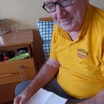 Na prvi fotografiji starejši gospod v rumeni majici z napisom "Poli Dog" sedi na postelji in v rokah drži ročno napisano pismo. Gospod se smehlja in nosi očala, v ozadju pa je omarica z osebnimi predmeti.
