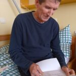 Četrta fotografija prikazuje moškega v temno modri majici, ki sedi na postelji in prebira pismo. Moški je osredotočen na vsebino pisma, v ozadju pa je vidna omara z nekaj predmeti in posteljno pregrinjalo s karo vzorcem.