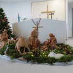 Prikazane so ročno izdelane jaslice z lesenimi figurami, obdane z mahom in okrašene z drobnimi lučkami. V ozadju sta okrašeno božično drevo in oltar z lesenim križem ter verskimi figurami.