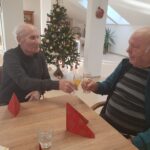 Dva starejša moška nazdravljata z napitki v prostorih, okrašenih z božičnim drevesom. Miza je okrašena z rdečimi servieti, ozadje pa poudarja praznično vzdušje z naravno svetlobo.
