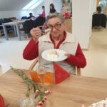 Starejša gospa sedi za mizo in z nasmeškom dviguje žlico proti kosu torte na krožniku. Na mizi so nageljni, kozarec s pijačo in rdeči praznični okraski. V ozadju se odvijajo praznične aktivnosti.