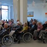 Občinstvo prikazano iz strani, kjer prevladujejo starejši poslušalci, mnogi na invalidskih vozičkih. Vidno je svetlo dnevno svetlobo skozi velika okna, dvorana pa je okrašena s snežinkami, ki visijo s stropa.