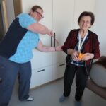 Zaposlena v domu za starejše s šibo v roki uprizarja šaljivo "tepežnico" z nasmejano starejšo gospo, ki stoji v sobi. Gospa v rokah drži darilo in z nasmehom pozira za fotografijo.