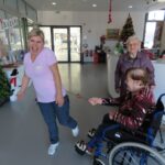 Na sliki starejša gospa v invalidskem vozičku šaljivo "tepe" zaposleno z okrašeno šibo. Obe se smejita in uživata v prazničnem vzdušju. V ozadju je božično drevo in praznični okraski.