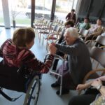 Na fotografiji dve starejši gospe sedita nasproti ena drugi, ena v invalidskem vozičku, druga s hoduljo. Veselo se držita za roke in se smejita. V ozadju so drugi stanovalci, ki spremljajo dogajanje v praznično okrašenem prostoru.