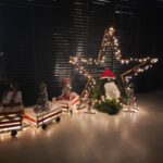 Na sliki je praznična dekoracija, postavljena v notranjem prostoru. V ospredju je okrasni vlak, narejen iz lesenih zabojev, okrašenih z lučkami, darilnimi škatlami in majhnim božičnim drevescem. Poleg vlaka stoji figura iz zelenega okrasnega materiala, okrašena kot Božiček, z rdečo kapo in belo brado. V ozadju je velika lesena zvezda, ovita z lučkami, ki ustvarja toplo in praznično vzdušje.