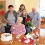 Starejša gospa, ki praznuje 90. rojstni dan, sedi za mizo ob torti, na kateri piše "Iskrene čestitke za 90 let!". Gospa v roza jopici drži kozarec in se nasmehne proti kameri. Ob njej stojijo štirje člani družine: starejši moški z belimi lasmi in brki, dve ženski, ena z rdečimi lasmi in druga z dolgimi svetlimi, ter še ena gospa z očali. Skupaj ustvarjajo prijetno vzdušje, obkroženi z rdečimi vrtnicami in slavnostno mizo.