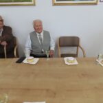 Za dolgo leseno mizo sedi starejši gospod v svetli telovnik z medaljo okrog vratu. Pred njim je krožnik s krofom, posut s sladkorjem, in kozarec penine. Ob njem sedi drug starejši gospod, oblečen v rjav pulover, ki drži palico. V ozadju na beli steni visijo slike z naravnimi motivi.
