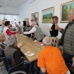 Skupina starejših ljudi stoji ob mizi in dviguje kozarce v zdravico. Na mizi so krofi in kozarci penine. Ljudje se smehljajo in voščijo slavljenki ali slavljencu. V ozadju so slike in bele stene, ki prostoru dajejo svetel videz.