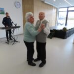 Starejši moški in ženska plešeta v prostoru z belo steno in velikimi okni, skozi katere se vidi zunanjost. V ozadju je glasbenik, ki igra na klaviature in poje. Na levi strani je videti še nekaj drugih ljudi, ki uživajo v glasbi in plesu.