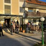 Skupina ljudi stoji pred vhodom doma za starejše. Nekateri so v invalidskih vozičkih, drugi pa stojijo na stopnicah ali na dvorišču. Stavba ima rumeno fasado, zunanjo teraso z lesenim ogrodjem in nadstreškom. Ob strani so okrasne rastline in majhna lesena hiška za ptičke. Vsi opazujejo praznično dogajanje.
