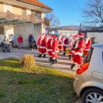 Pred stavbo stoji skupina ljudi, oblečenih v kostume Božičkov. Nekateri nosijo harmoniko in druge instrumente. Na desni strani slike je parkirano osebno vozilo, v ozadju pa so vidni prebivalci doma za starejše, ki opazujejo dogajanje. Prostor je sončen, z nekaj zimskega zelenja in brez listja na drevesih.
