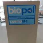 Na sliki je naprava z imenom "biopol +6", ki je povezana z bioenergijo za izboljšanje počutja. Na napravi je logo "cosmolife" in napis "Better Life Products". Izdelek je nameščen v notranjem prostoru, ozadje pa kaže del prostorov, vendar ni popolnoma jasno vidno. Naprava je svetlo sive barve.
