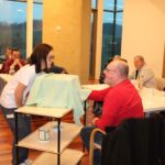 V svetli sobi, kjer so na levi strani velika okna z razgledom na podeželsko pokrajino, ženska v beli majici kleči ob premični mizi, prekriti z zelenim blagom. Nasproti nje sedi moški v rdečem puloverju, ki z nasmehom sodeluje pri dejavnosti. Za njima sedijo še drugi udeleženci za mizami, nekateri so nasmejani in spremljajo dogajanje. Na tleh so lesena tla, ki dodajajo toplino prostoru.